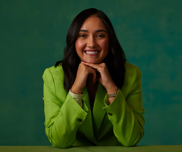 Yaritza Vallejo poses for a photo, resting her chin on her hands and smiling. She wears a light green blouse and sits in front of a dark green background.