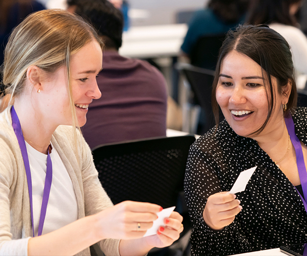 Two women, both Kellogg MBA students, laugh and smile as they participate in a conversation and show each other notecards. Both are wearing purple lanyards.